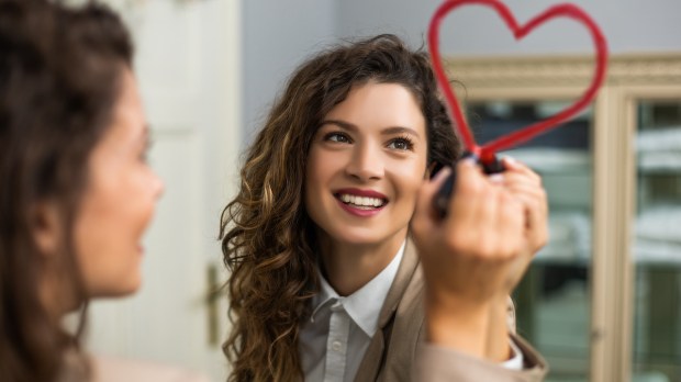 Businesswoman is drawing heart with lipstick on the mirror while preparing for work