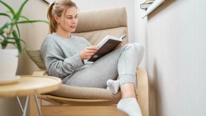 Blonde woman reading book while sitting on beige armchair in apartment