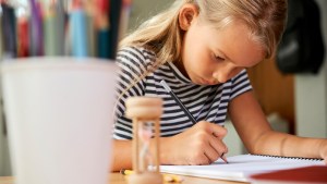 Pretty little diligent schoolgirl with hourglass on her desk doing homework