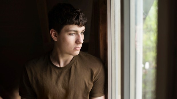 A young teenager boy with sadness in his eyes looks out the window