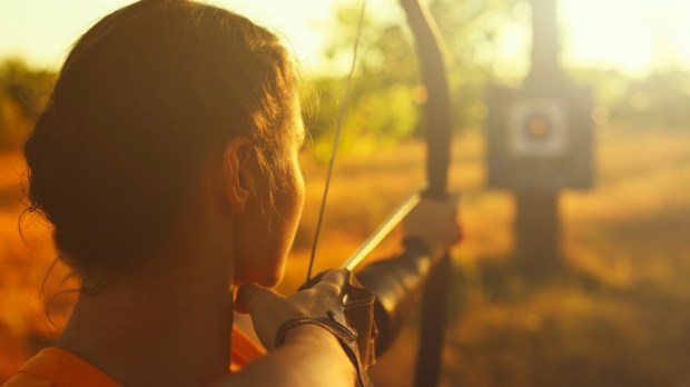 female archer shooting with a bow in a field at sunset