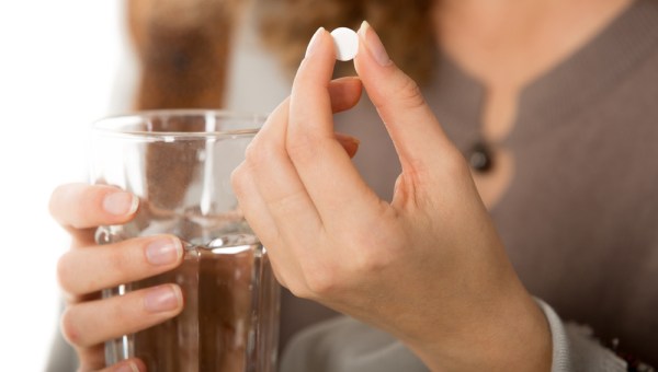 female hands holding one white round pill and glass of water