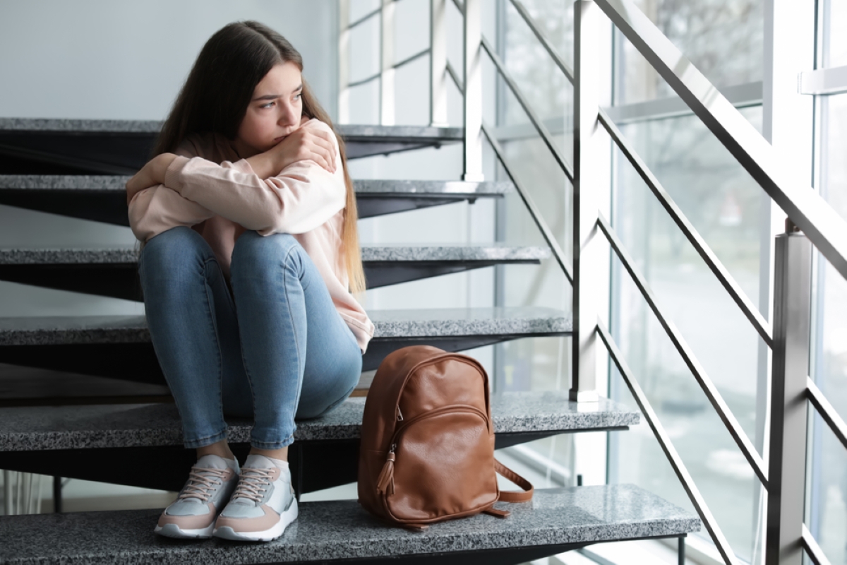 Upset teenage girl with backpack sitting on stairs indoors