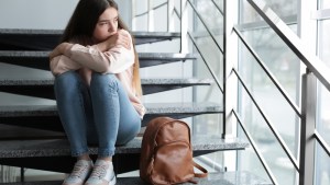 Upset teenage girl with backpack sitting on stairs indoors