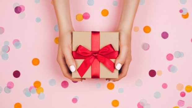 Woman hands holding present box with red bow on pastel pink background with multicolored confetti