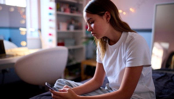 Depressed Teenage Girl Sitting On Bed At Home Looking At Mobile Phone