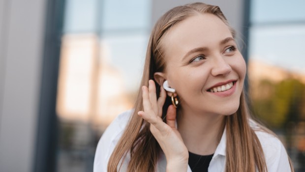 Happy blonde woman sitting on the bench near office building with wireless earbuds in ears
