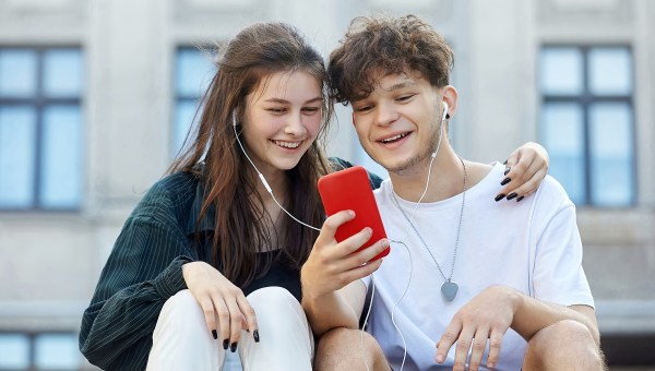 young guy and a girl sitting on the street, who have one pair of headphones for two, look into a red smartphone with a smile.