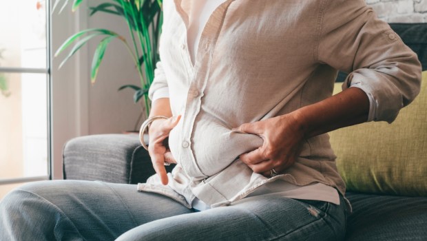Midsection of woman hands holding her belly fat sitting on sofa at home.