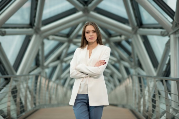 a young brunette girl in a white jacket is standing in a bright corridor of tubular metal structures looking at the camera